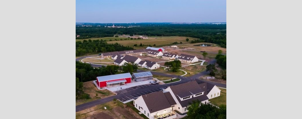 Aerial view of several low-rise residential buildings, a barn, and a greenhouse with fields and forests in the background.