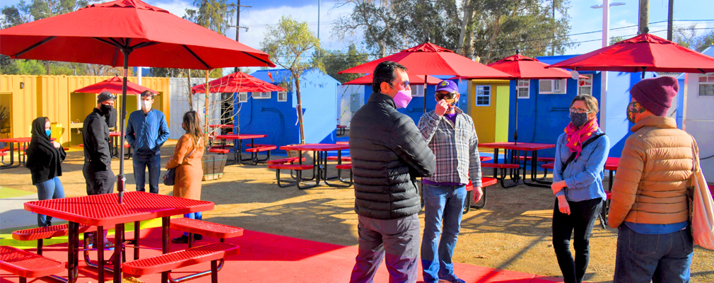 Photo of people gathered at an area with red outdoor tables with red umbrellas with colorful tiny homes in the background.