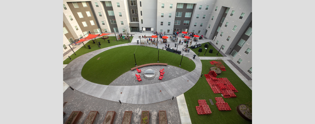 Photograph taken from an upper story of a courtyard with a fire pit, raised garden beds, artificial turf, and exhibit tables and umbrellas for a community gathering.