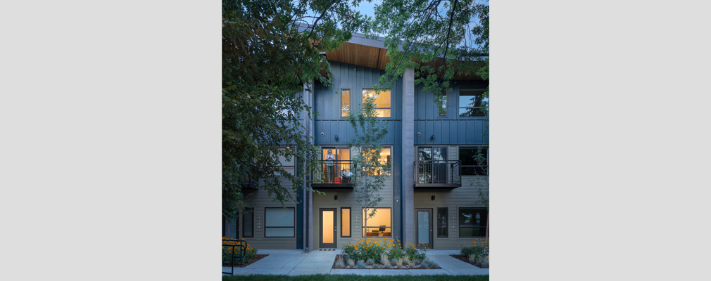 Three attached residences, with a woman standing on the second-story balcony of the middle unit and landscaping in the foreground.