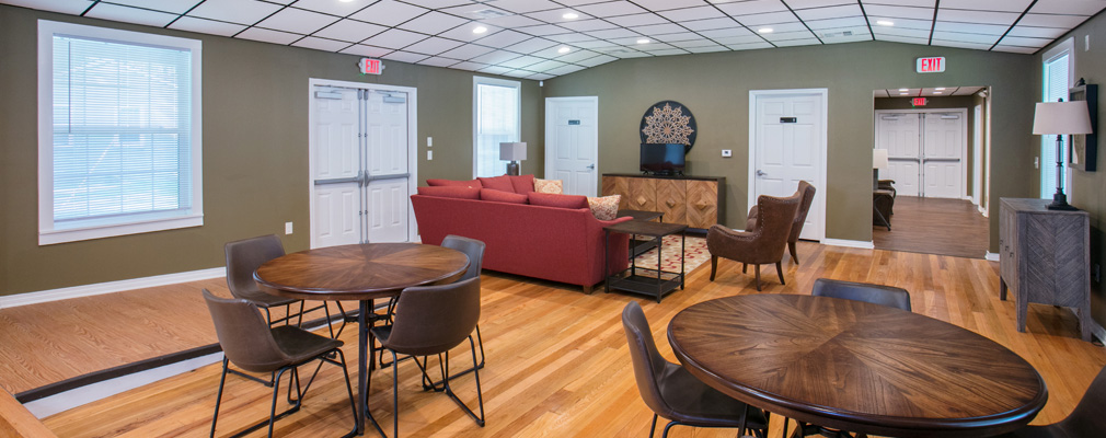View of the community room in Harmony Village, with tables and chairs in the foreground and a couch and cabinets in the background.