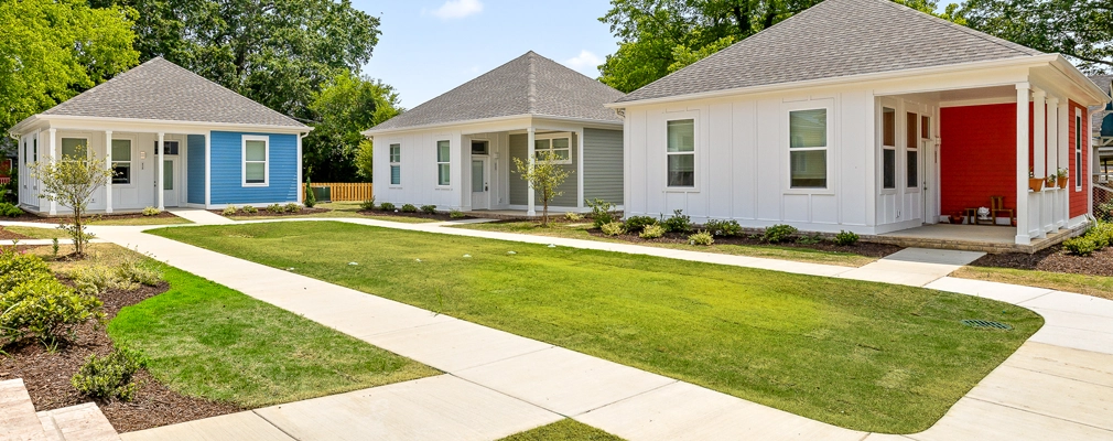 A perspective photograph of three single-story cottages with covered front porches arranged around a central lawn with curved concrete walkways and landscaped planting beds.