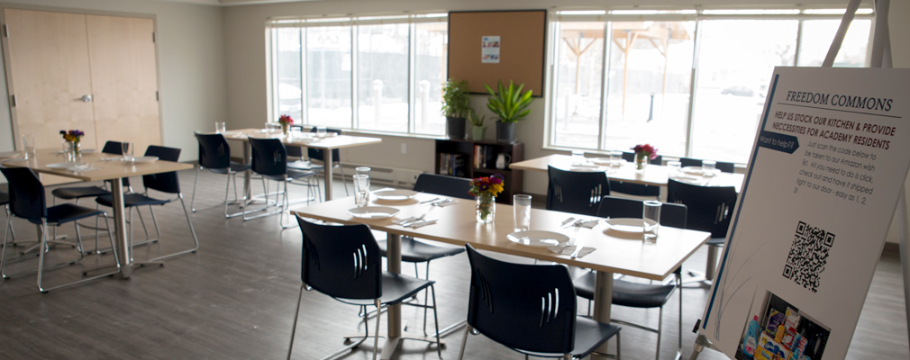 Photograph of a large room with four tables set with dinnerware, large windows in the rear wall, and a poster on an easel at the right edge of the photograph. 