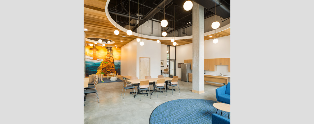 Interior photograph of a room with a ceiling with exposed ductwork, a table with chairs, and a kitchenette, with a children’s play space framed by a mural of a totem pole in the background.