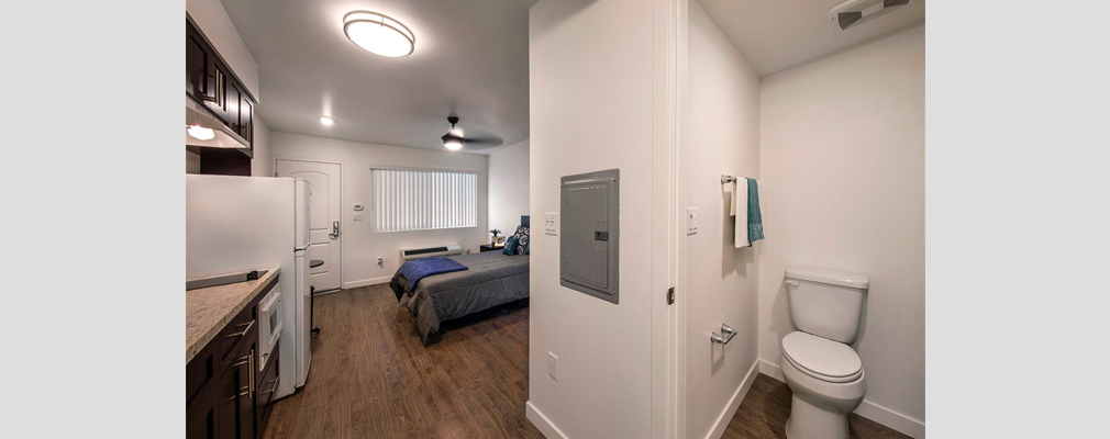 Photograph of apartment unit interior with a bathroom, bed, and kitchen cabinets.