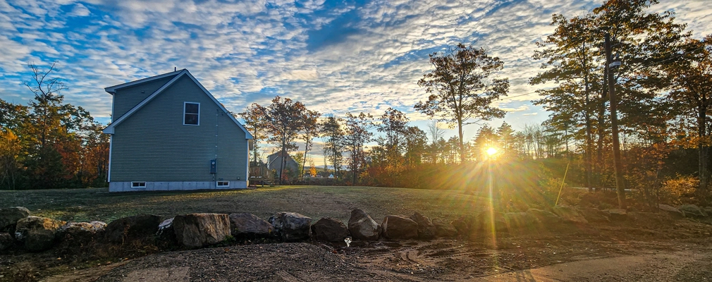 Side view of a two-story house in a rural area with trees and a sunrise in the background. 