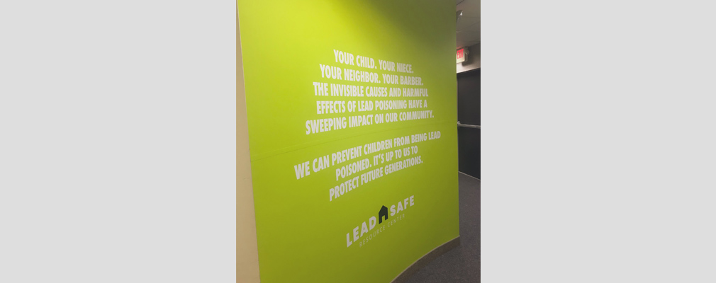 Photograph of a green-painted graphic wall that reads ”Your child. Your niece. Your neighbor. Your barber. The invisible causes and harmful effects of lead poisoning have a sweeping impact on our community. We can prevent children from being lead poisoned. It’s up to us to protect future generations.”