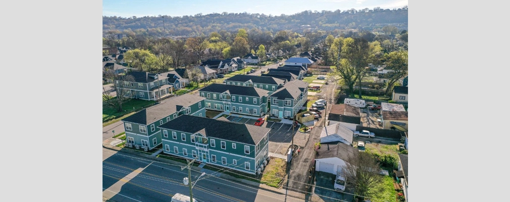 An aerial photograph of multiple two-story buildings with pitched roofs, in a grid layout with parking areas, and a hilly landscape in the background.