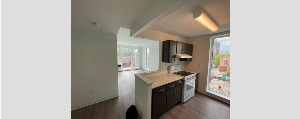 Image of an empty apartment unit's kitchen and living room space with floor-to-ceiling windows.