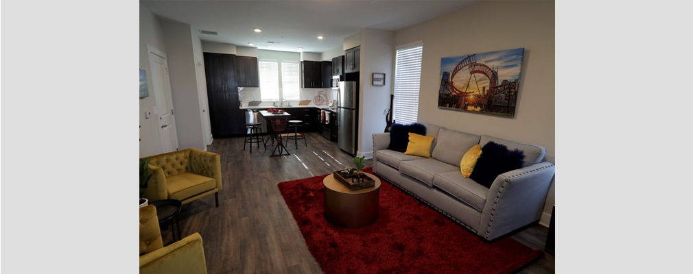 Photograph of a living room with a couch and two chairs in the foreground and a kitchen with a high table and stools in the background. 