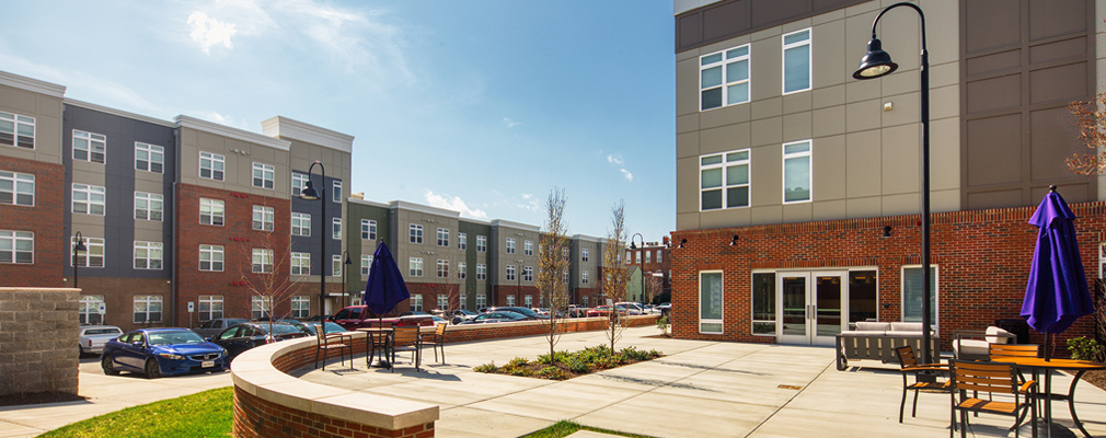 Photograph of a patio in front of the side façade of a three-story residential building (Van de Vyver) and the rear façade of a four-story residential building (the Rosa), with a parking area between the buildings.