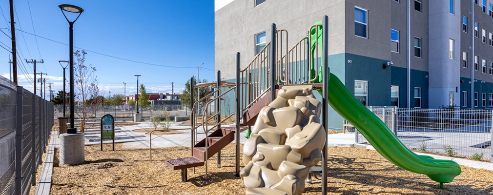 An exterior photograph of a playground with a slide and rock-climbing feature surrounded by a fence and bordered by a four-story building.