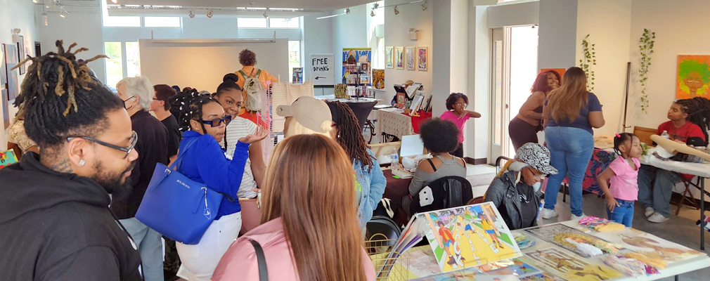 Room with people gathered and art displayed on tables and walls.