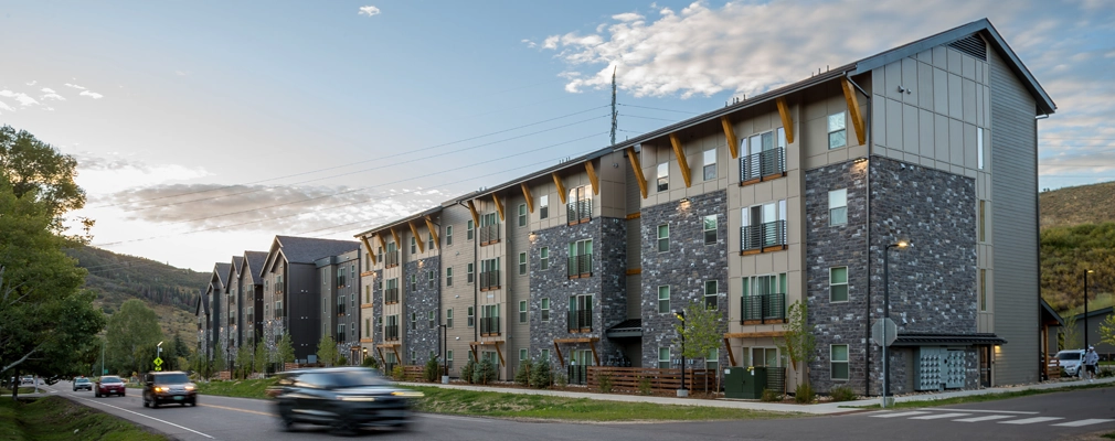 A perspective street-level view of a four-story building with cars driving past on the road and hills in the background.