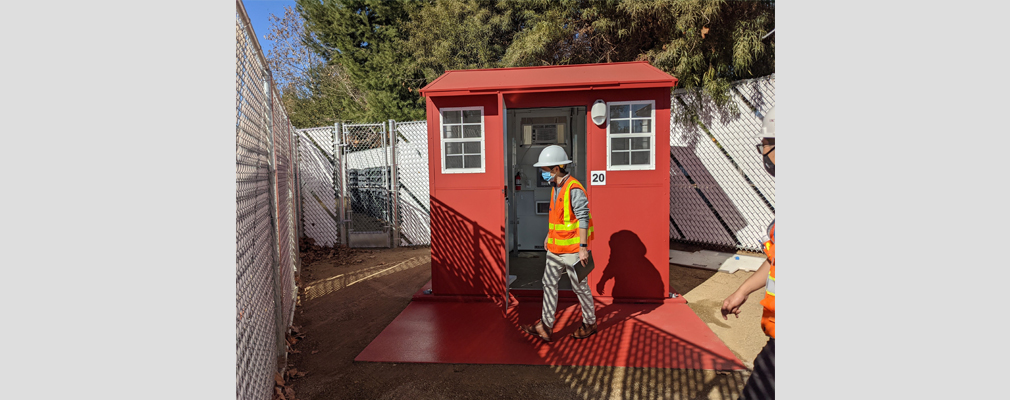 A photograph of a red tiny home with the door open, revealing a white interior with a person in a hardhat and safety vest walking across the front.