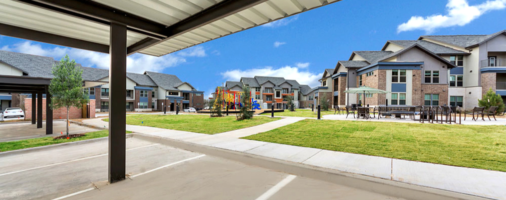 An apartment complex with a carport in the foreground, common areas in the middle-ground, and multistory residential buildings in the background.