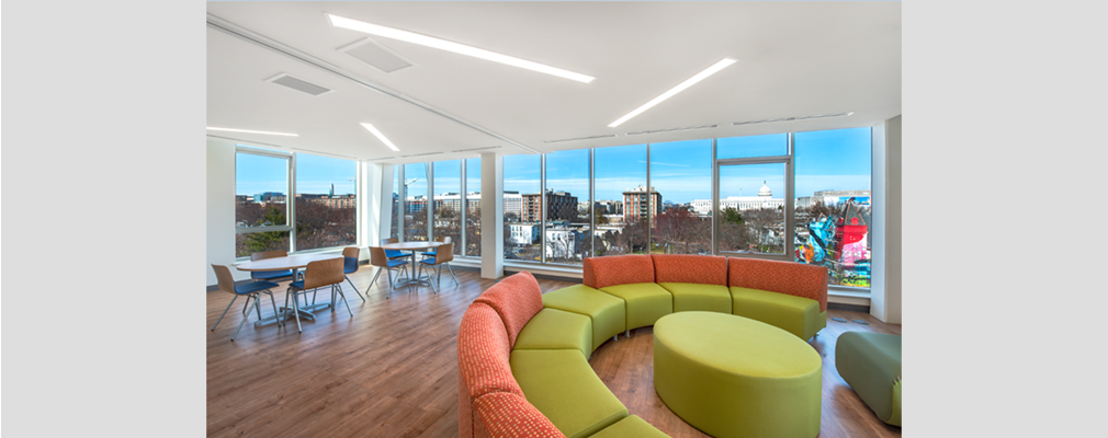 Large community room with seating areas and tall windows on multiple sides providing views of the city, including the U.S. Capitol building.