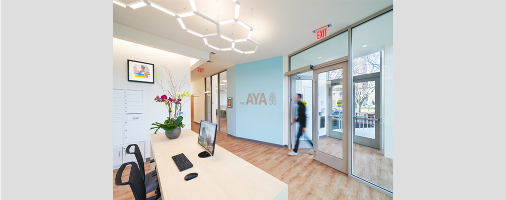 A reception area with desk and chairs facing glass entry doors and "The AYA" printed on one wall. 