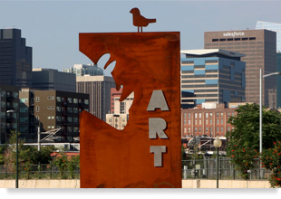 Photograph of an upright iron plate with carvings of a rhinoceros head and a bird and the word “ART” and with the skyline of downtown Denver in the background.