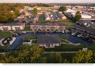 An aerial drone photograph of a central single-story building surrounded by a parking lot with cars and green space, with several two-story buildings in the background.