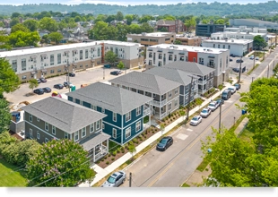 A low aerial photograph of a row of 5 two-story quadplexes with pitched roofs and front porches, fronting a residential street with parked cars. Several three-story apartment buildings are in the background.