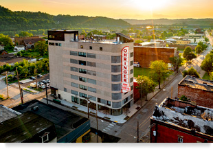 Low-angle aerial photograph of an eight-story building with a five-story blade sign that reads "Ohringer Home Furniture" in the foreground and the Monongahela River and mountains in the background.
