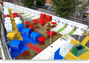 An aerial photograph of a brightly painted tiny home village surrounded by a white chain-link fence and white-painted highway barriers.