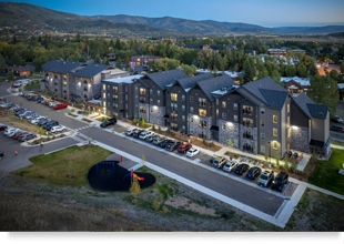 An aerial view of a four-story building surrounded by parked cars in front, a playground in the foreground, and mountains in the background.