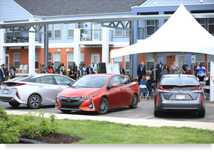 Photograph of the front façade of a two-story residential building, with a crowd gathered beneath a temporary pavilion and three hybrid vehicles on display in the foreground. 
