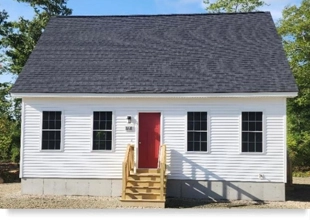 Front view of a small single-family house with white siding, a red door, and several windows. Source MaineHousing.