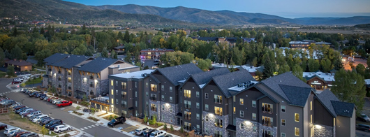 An aerial view of a four-story building surrounded by parked cars in front, a playground in the foreground, and mountains in the background.