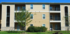 An apartment complex with a lawn, small trees and shrubbery, and a picnic table in front.