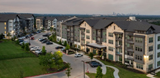 An aerial photograph of an apartment complex of multiple four-story buildings connected by a road lined with parked cars, surrounded by green lawns and a city skyline in the background.