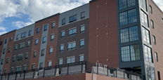 A modern red brick apartment building stands on a residential street. An older-looking three-story building is visible next to it.