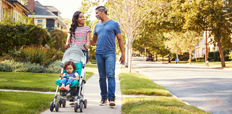 A couple walks down a sidewalk in a neighborhood while pushing a stroller with a child.