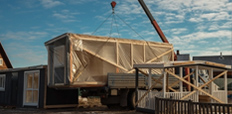 A crane hoists a house on a truck, illustrating the transportation of a building structure.