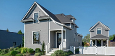 A gray house with a driveway, and a smaller house next to it and behind a white fence.