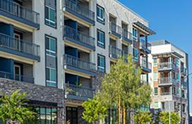 Modern multi-story apartment buildings with balconies with trees and greenery in the foreground under a clear blue sky.
