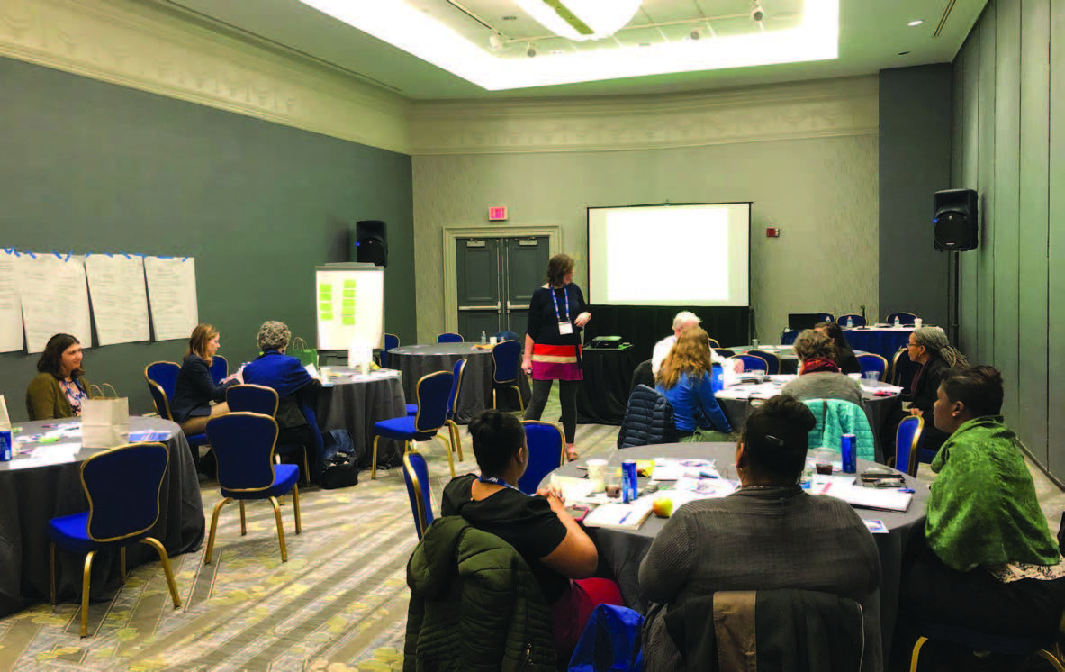 People seated at round tables in a room listening to a presentation by an individual in front of a projector screen.