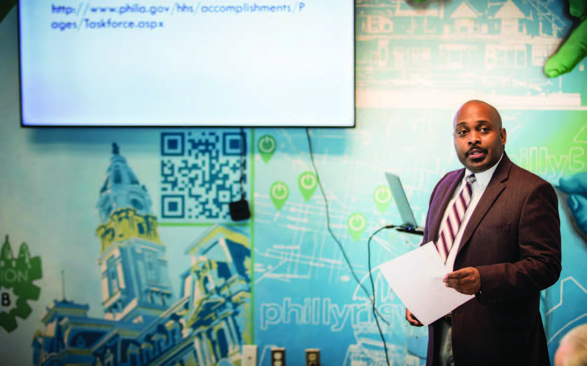 An individual standing in front of a mural wall with a television screen giving a presentation.