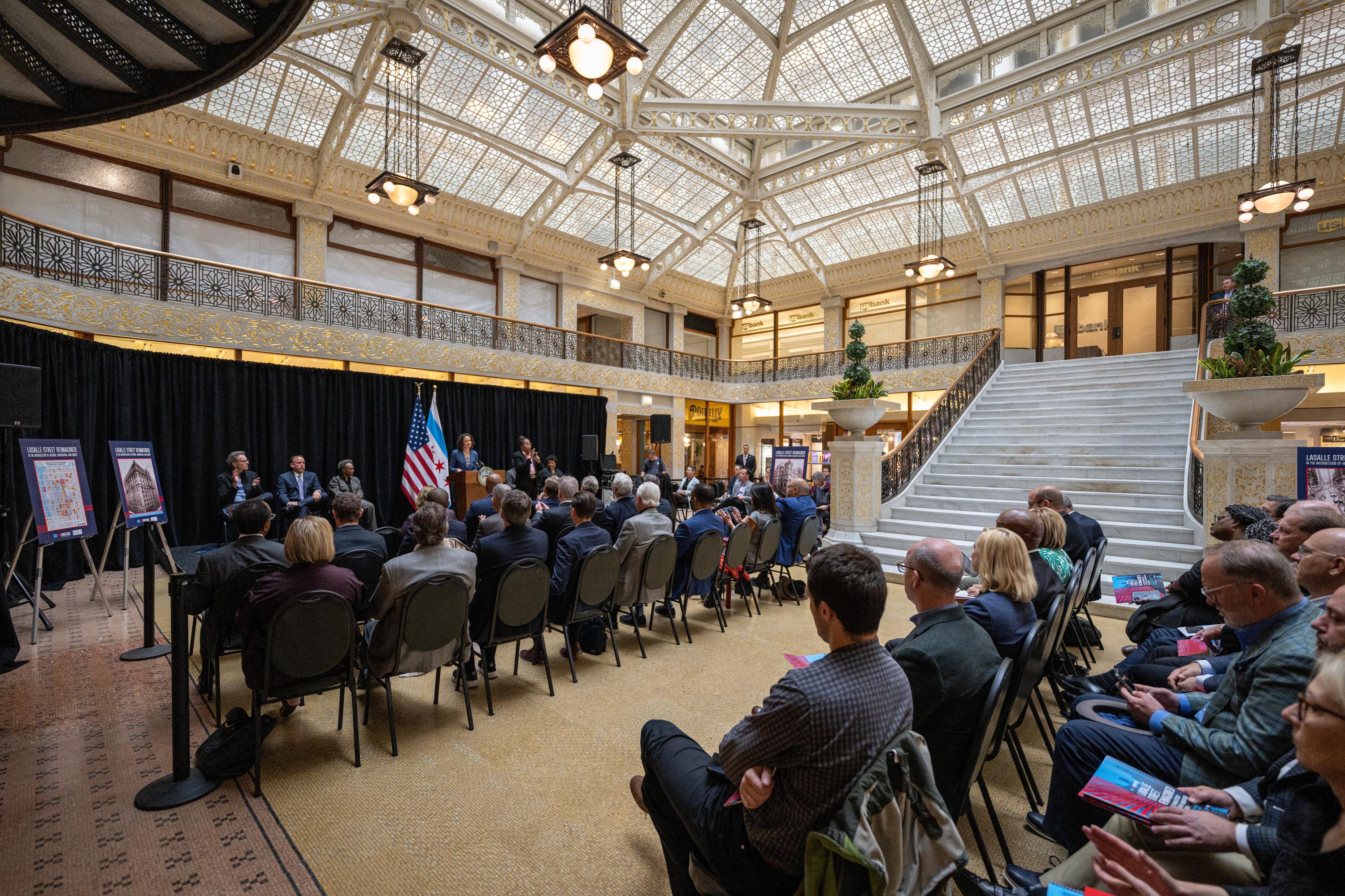 Audience seated in multiple rows listening to a presentation in a large room with a framed glass roof.