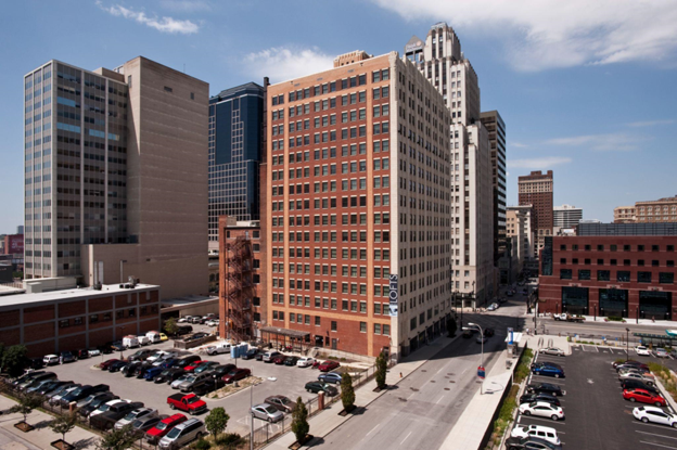 A tall multistory building with a parking lot in the foreground and other multistory buildings around it.