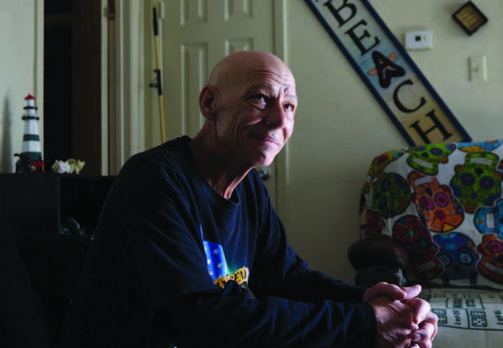 A seated man in a room with a sofa seat next to him and the sign “Beach” on the wall in the background.