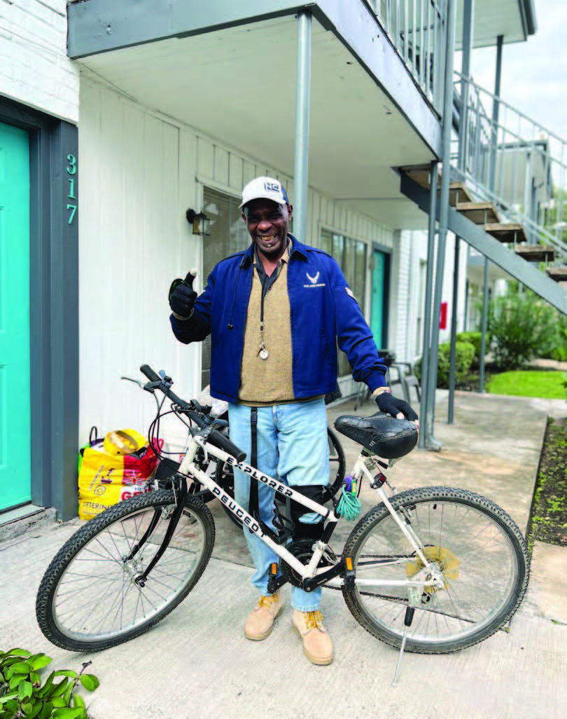 A man standing and holding his bicycle in front of a door of a two-story residential building.