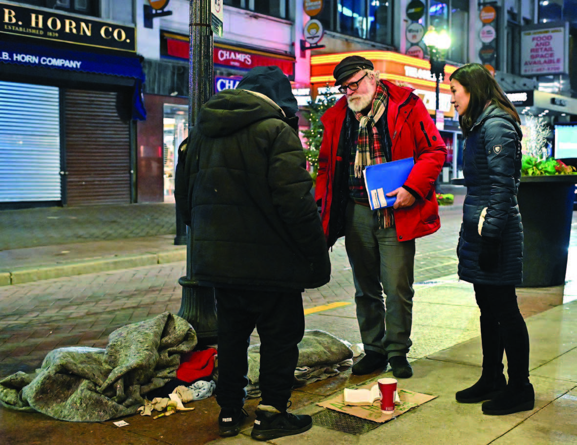 Three people standing on a sidewalk with store fronts on the other side of the street.