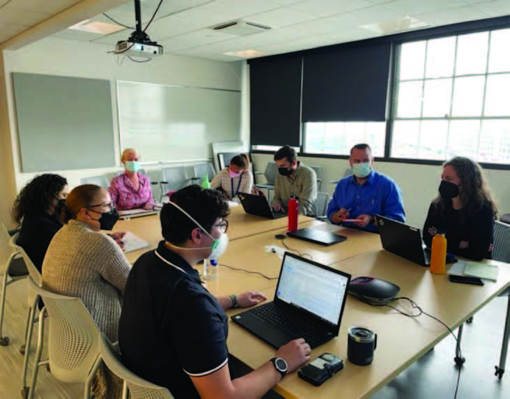 A group of eight people seated around a large table with a projector on the ceiling and a white board on one wall.