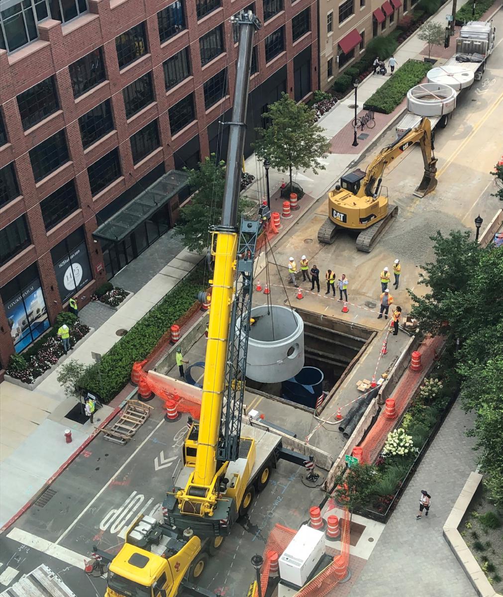 Photo of a crane lowering stormwater equipment into a hole in the middle of a street with multistory buildings on one side.
