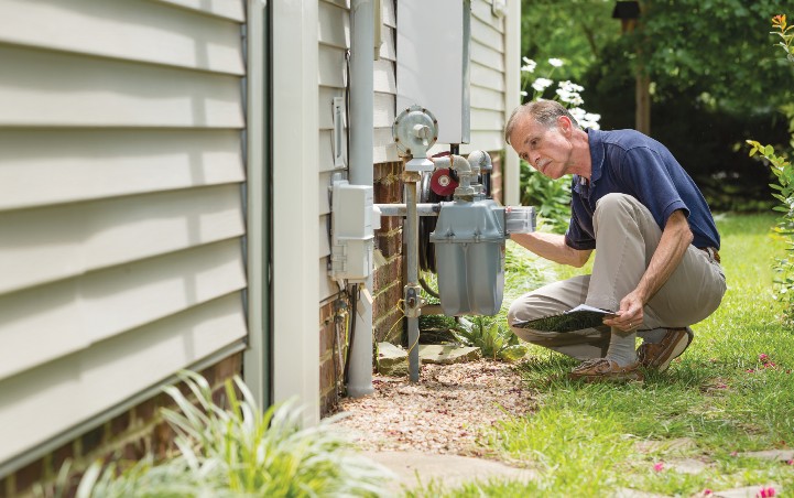 A man kneeling down to study a meter on the side of a house.