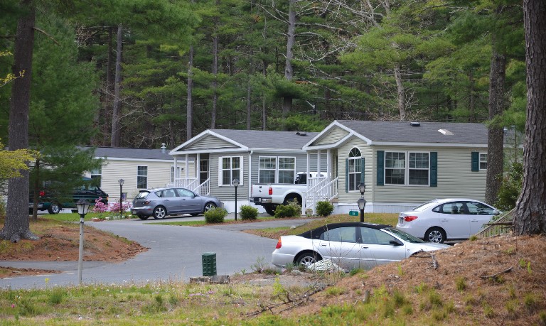 A curved street with three manufactured homes with a densely wooded background.