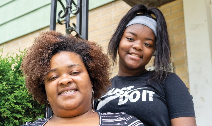 A close-up of a woman and a girl in front of a siding and brick background and a scroll design black wrought iron column.